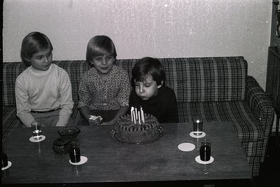 Three children in 1970s-style clothing gather around a birthday cake with lit candles on a round table. Plaid sofa and patter...