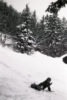 Snowboarder mid-fall on groomed slope, surrounded by dense coniferous forest. Classic 1990s winter sports attire—helmet, gogg...
