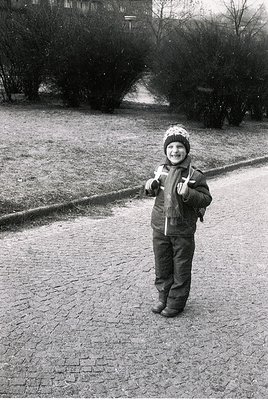 Young child in winter attire—knit hat, padded coat, and boots—stands on a cobblestone path in a park-like setting. Snowy tree...