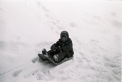 Child sledding on snow-covered slope, mid-20th century winter attire. Black-and-white photo captures joyful winter activity i...