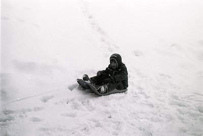 Black-and-white snapshot of a child sledding on a snow-covered slope, mid-20th century. The child wears a hooded jacket and s...