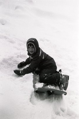 Child sledding on snow-covered ground, wearing a striped knit hat, dark winter coat, and mittens. Classic winter activity, li...