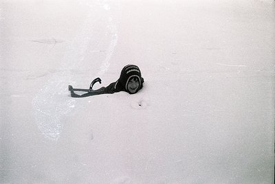 Vintage analog camera partially buried in snow, strap coiled. Minimalist composition highlights contrast between cold white a...