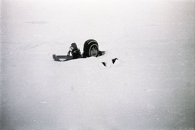Black-and-white close-up of a vintage camera lens partially buried in snow, likely a **Leica-style rangefinder**. The composi...