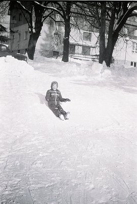 Child sliding down snow-covered hill in urban park, mid-1970s. Snowy terrain contrasts with bare winter trees and multi-story...