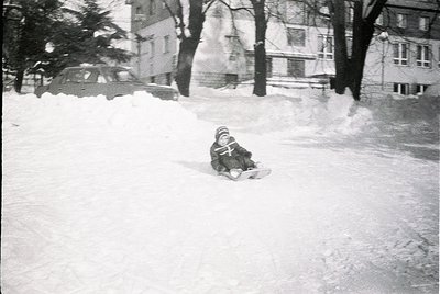 Child sledding on snow-covered urban slope, mid-20th century. Snow piles and parked vintage car in background suggest residen...
