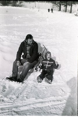 A man and child sledding in snow, mid-20th century. The man, seated, guides a child on a sled, both dressed in winter attire—...
