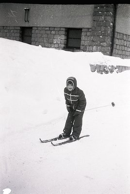 Child in vintage ski gear poses on groomed slope beside brick building, mid-20th century. Snow-covered terrain and classic sk...