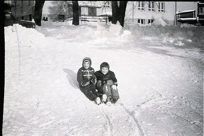 Two children sledding on a snow-covered street in mid-20th century urban setting. Snow piles and parked cars suggest resident...
