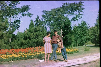 Vintage color photo of three individuals in a lush park setting, likely 1960s–1970s. Woman in floral dress, man in plaid shir...