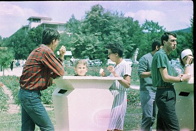Vintage group photo in a landscaped park setting, likely 1970s–1980s. Four adults and a child pose near a concrete bench surr...