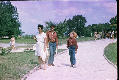 Vintage 1960s-70s park scene: woman in sleeveless dress, man in plaid shirt/jeans, and child in plaid shirt/jeans walking on ...