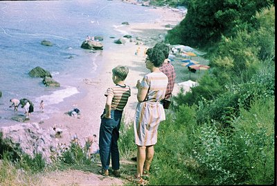 A mid-20th century seaside scene: two adults and a child stand on a grassy cliff overlooking a rocky shoreline. The woman wea...