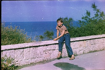 Vintage seaside shot of a boy in 1960s-style striped shirt and jeans leaning on a stone wall, overlooking the ocean. Warm sep...