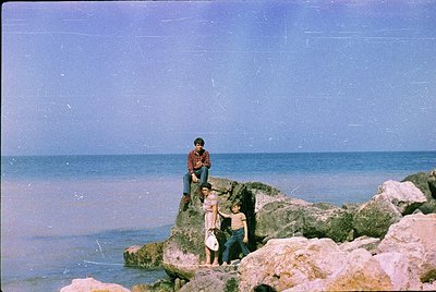 Vintage seaside group shot on rocky coastline, likely 1970s–1980s. Four individuals pose on weathered rocks near calm waters,...