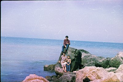 Vintage seaside family portrait on rugged rocks, likely Mediterranean. Three individuals pose on uneven stone formations near...