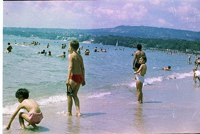 Crowded seaside beach scene with children playing in shallow waves, 1960s-70s. Foreground shows three boys in swim trunks—one...