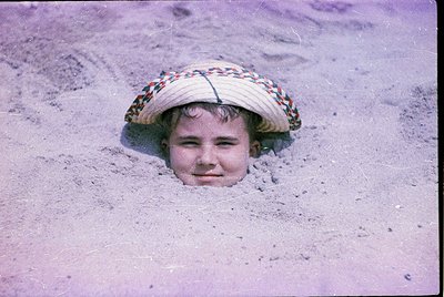 Child peeking from a sand tunnel, wearing a wide-brimmed straw hat with colorful woven trim. Mid-20th century beach scene, li...