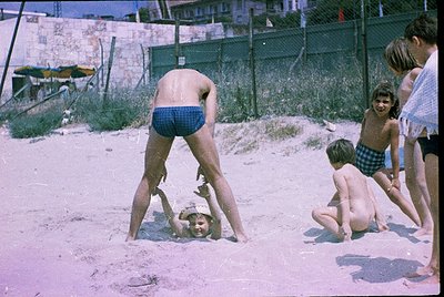 Vintage beach scene with children playing in shallow water, mid-20th century. A shirtless boy in plaid shorts lifts another c...