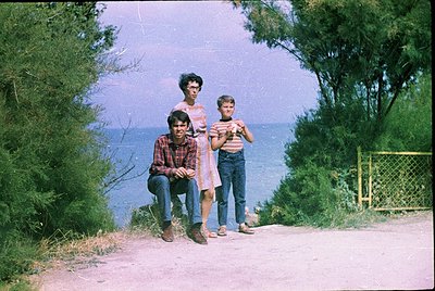 Family portrait by a seaside cliff, 1970s. Three individuals pose on a gravel path with ocean backdrop, framed by greenery. T...