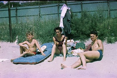 Vintage beach scene featuring three individuals lounging on a sandy shore beside surfboards. The adult on left wears red shor...