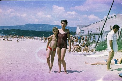 Vintage seaside scene from mid-20th century, likely 1950s–60s. Woman in patterned one-piece swimsuit poses with young boy in ...