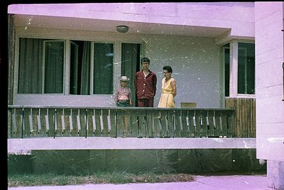 Vintage 1960s family portrait on a mid-century porch with white vinyl siding. Three individuals pose: a child in a striped sh...