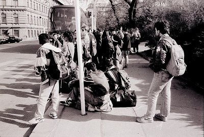 Black-and-white street scene featuring a group of young people waiting at a bus stop in an urban setting. One person sits on ...