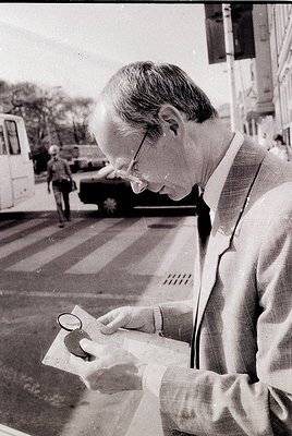 Mid-20th century man examines a map with a magnifying glass on a city street, likely 1950s–1960s. Urban setting with parked v...