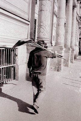 Mid-20th century street scene: Man in mid-stride under ornate umbrella, holding a second open umbrella. Architectural details...