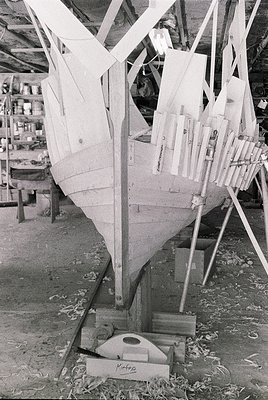 Traditional wooden boat under construction in a dry dock, featuring ribbed frames and planks. The hull’s curved shape suggest...
