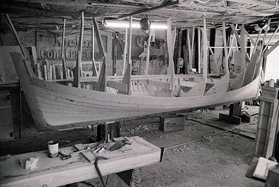 Traditional wooden boat under construction in a workshop, framed ribs visible. Sign reads "Kiel Construction." Mid-20th centu...