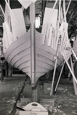 Half-built wooden boat hull in a dry dock, secured by metal supports and planks. Traditional planking and rib construction vi...