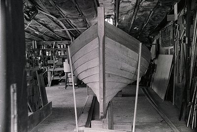 Mid-20th century workshop showcasing traditional wooden boat construction. Planks and ribs form the hull of a small vessel, s...