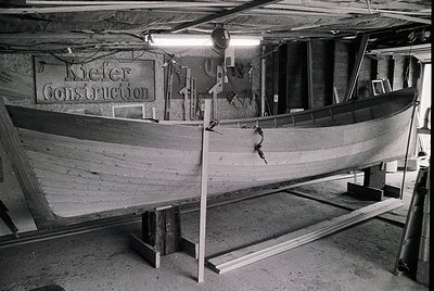 Industrial boat construction scene inside a workshop labeled "Kiefer Construction." A wooden boat hull rests on supports, wit...