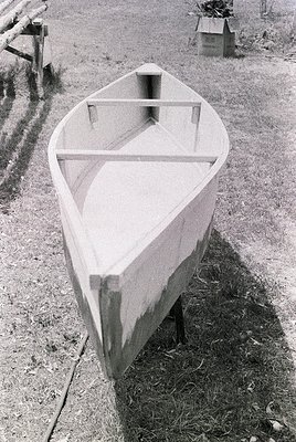 Classic wooden rowboat with simple crossbeam seating, resting on dry grass near a rural path. Likely mid-20th century design,...