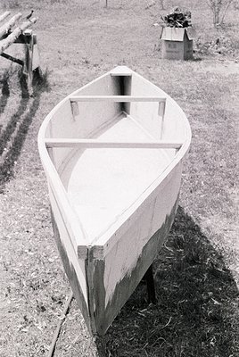 Classic wooden rowboat with simple frame structure, resting on grassy ground near a dirt path. Rustic wooden fence and small ...