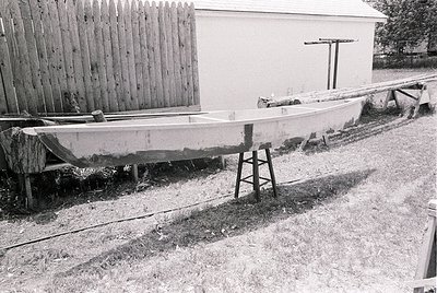 Vintage wooden boat resting on a makeshift cradle beside a weathered wooden fence and small shed. Rustic rural setting, likel...