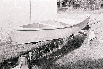Vintage wooden boat resting on makeshift supports in a grassy area, likely mid-construction or repair. Visible planks, nails,...