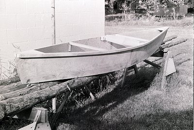 Classic wooden rowboat resting on makeshift wooden supports in a grassy yard. Mid-20th century construction with smooth, roun...