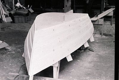 Mid-century industrial boat hull in fiberglass construction, draped in protective cover. Factory setting with wooden molds, t...
