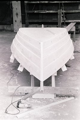 Mid-stage wooden boat hull construction in a workshop, featuring plank-on-bulkhead framing. Power tool and wooden supports vi...