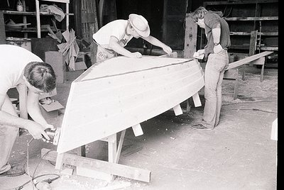 Three men collaborate on assembling a wooden structure in an industrial workshop, likely mid-20th century. Workers wear caps,...