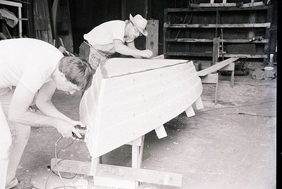 Two men in 1950s-60s workwear sand and shape a large wooden boat hull in an industrial workshop. Visible tools include sandpa...