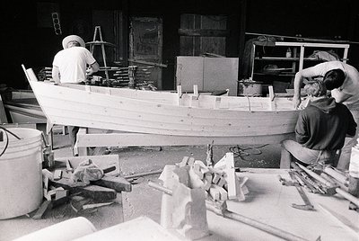 Two craftsmen build a wooden boat in a workshop, surrounded by tools and materials. Mid-20th century industrial craftsmanship...