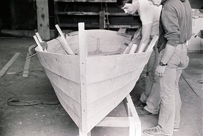 Handcrafted wooden boat under construction, likely mid-20th century. Two workers inspect planks and ribs in an industrial wor...
