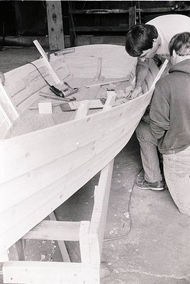 Traditional wooden boat construction in progress, featuring two craftsmen shaping hull planks with hand tools. Industrial set...
