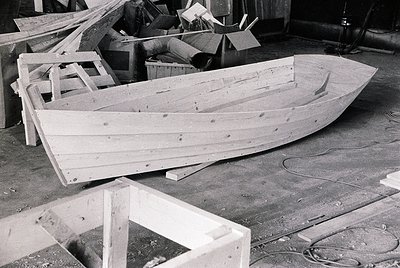 Unfinished wooden boat frame in a workshop, likely mid-construction. Ribbed hull with planks secured by nails, surrounded by ...