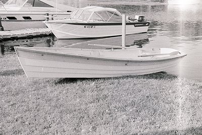 Classic wooden sailboat on grassy shore, mid-20th century design. Foreground boat features simple hull, transom stern, and mi...