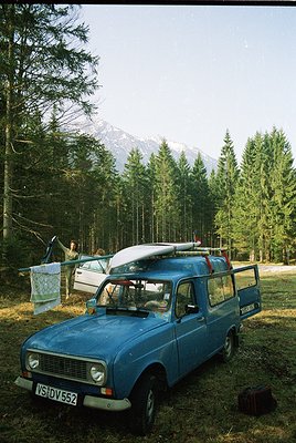 Vintage blue station wagon (likely 1970s) parked in a forested area, carrying surfboards on its roof. A person stands near a ...